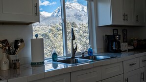 A kitchen with white cabinets, a window with a view of snow-capped mountains, a sink, and various kitchen items. - Skyview Terrace (Saguache County)