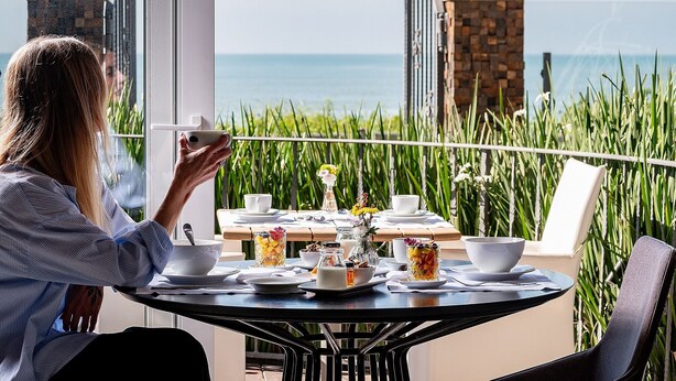 A person at a table with a view of the sea, a balcony with a railing, and a table set with dishes and a glass of fruit. - Villa d'Ozio Hotel e Cucina (Itajai)