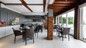 A modern dining area with a high ceiling, exposed wooden beams, and large windows offering a view of greenery outside. - Villa d'Ozio Hotel e Cucina (Itajai)