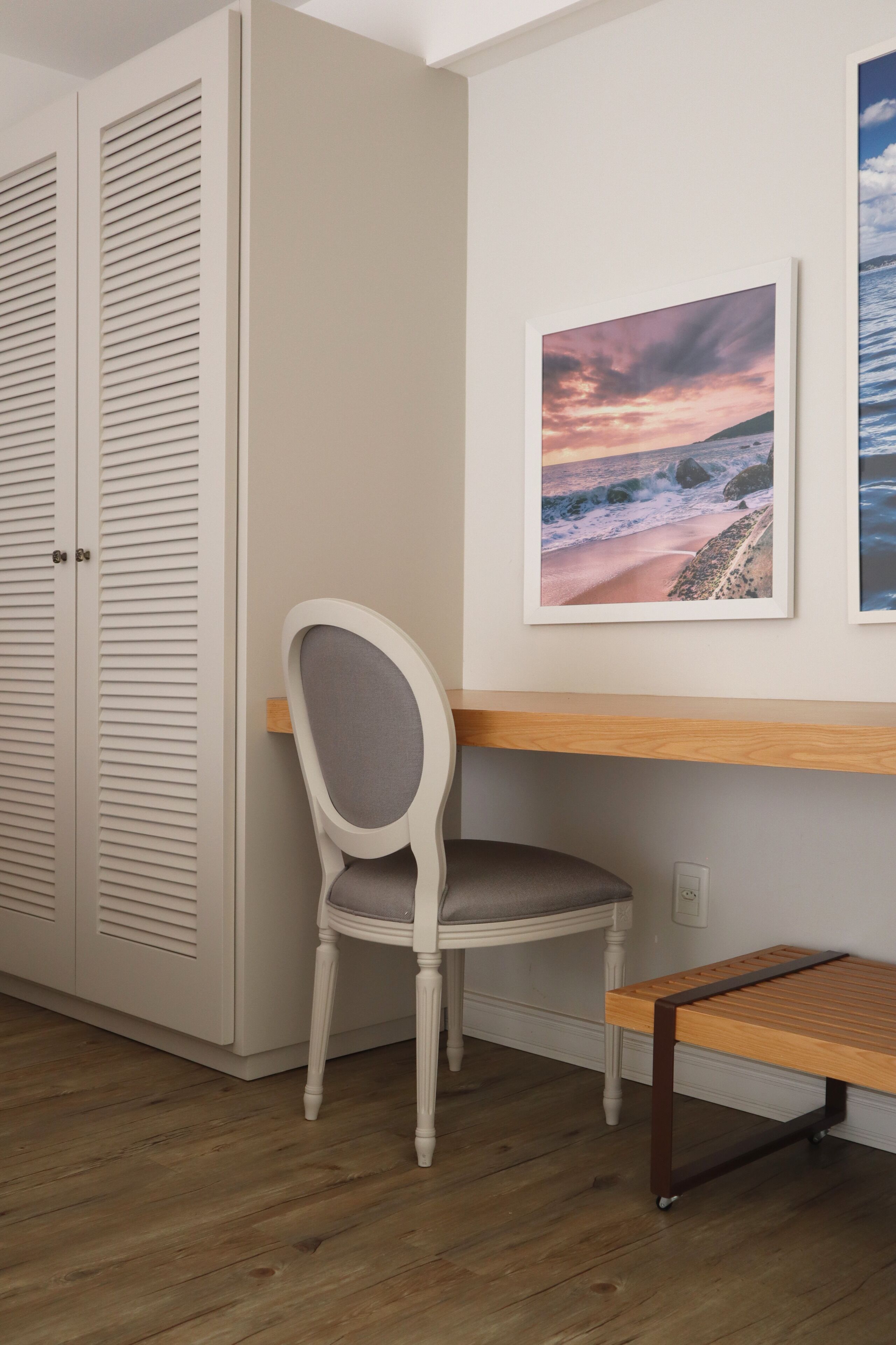 Una habitación con suelo de madera, una silla blanca, una mesa de madera, un armario blanco y dos cuadros en la pared.