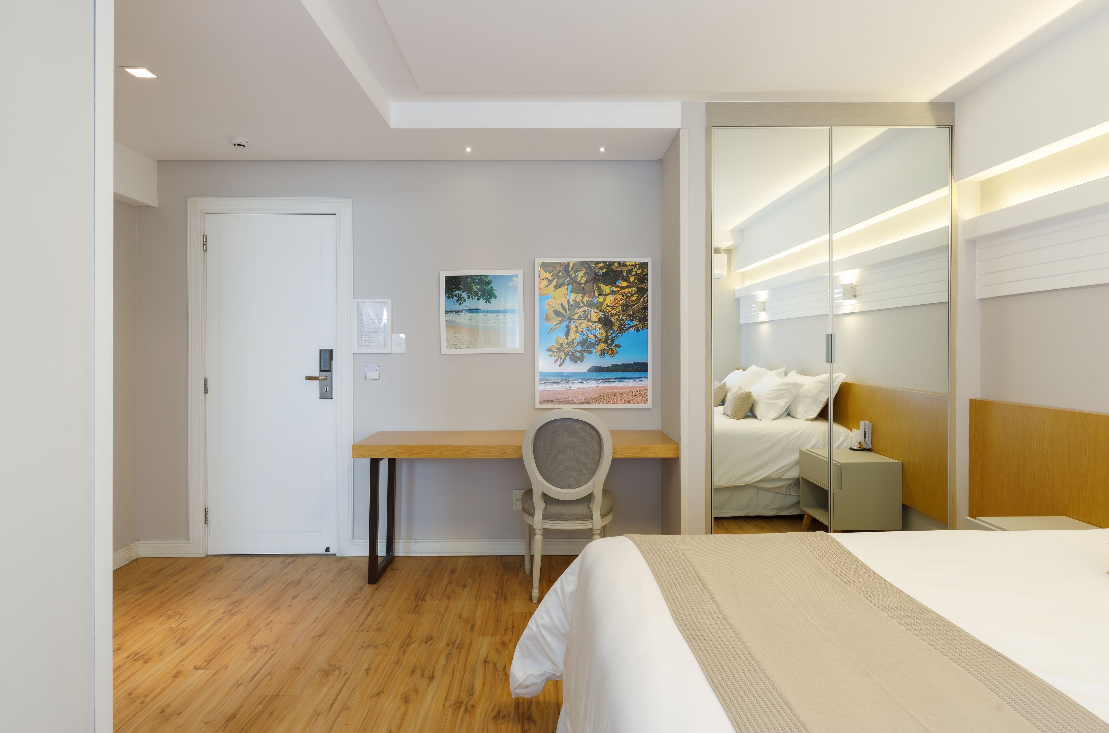 A hotel room with a bed, a desk, and a chair. There is a door, a wall with a painting, and a mirror.
