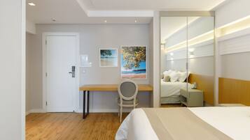 A hotel room with a bed, a desk, and a chair. There is a door, a wall with a painting, and a mirror.