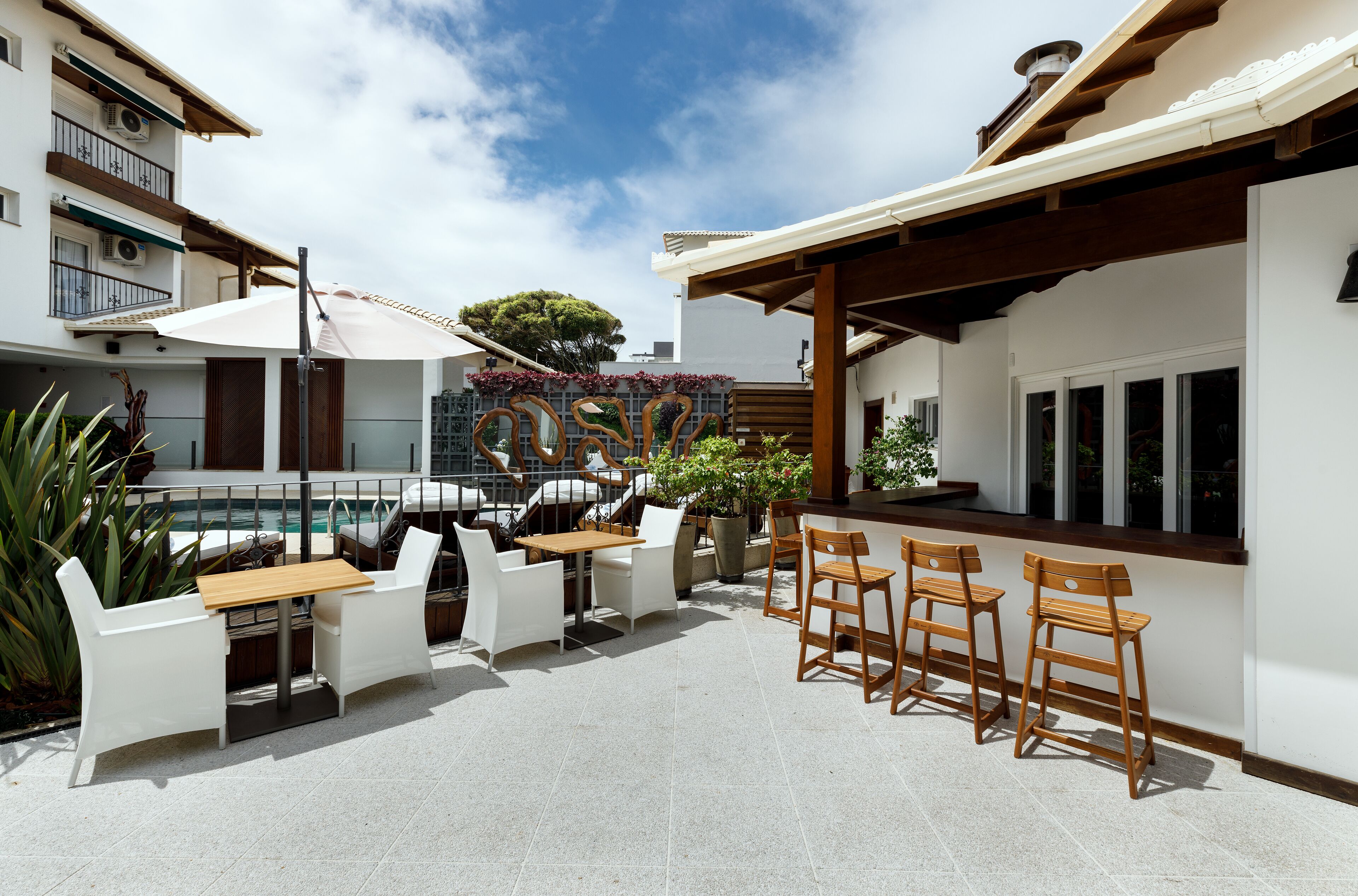 An outdoor patio with a pool, white chairs, wooden tables, and a bar area.