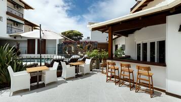 An outdoor patio with a pool, white chairs, wooden tables, and a bar area.