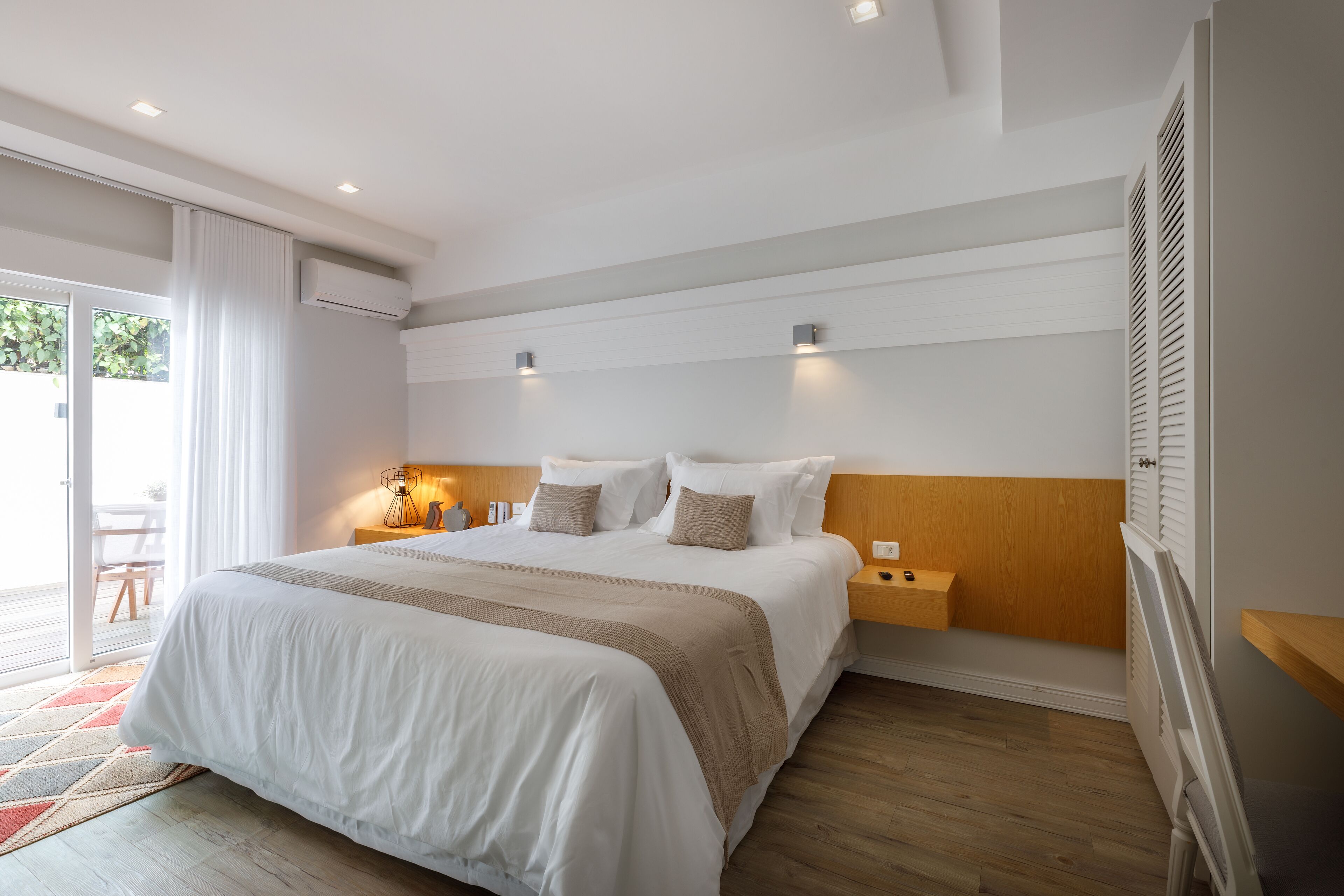 A modern hotel room with a large bed, wooden headboard, bedside tables, and a sliding glass door to the balcony.