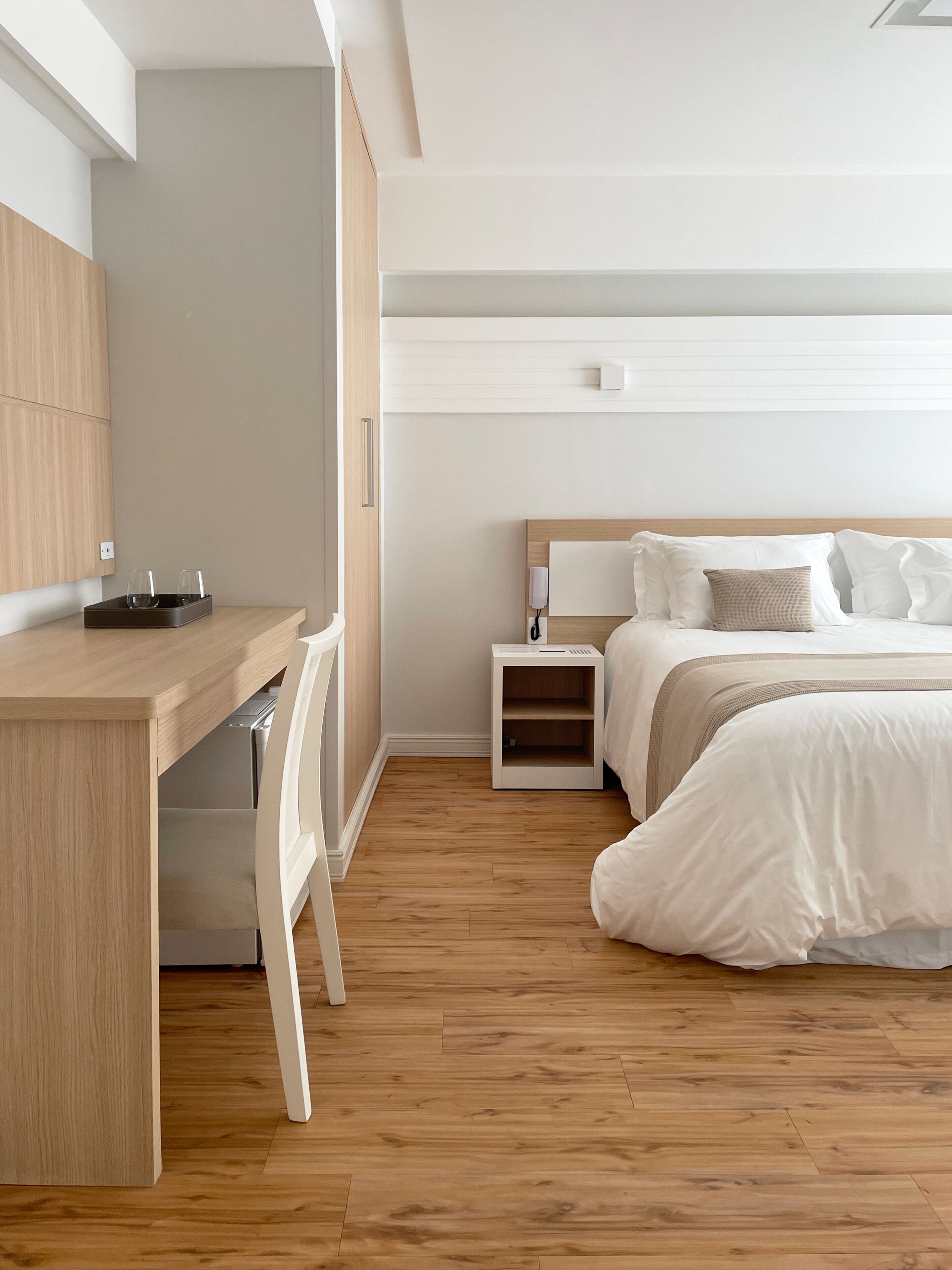 A modern bedroom with a wooden desk, a bed with white bedding, and a bedside table with a lamp.