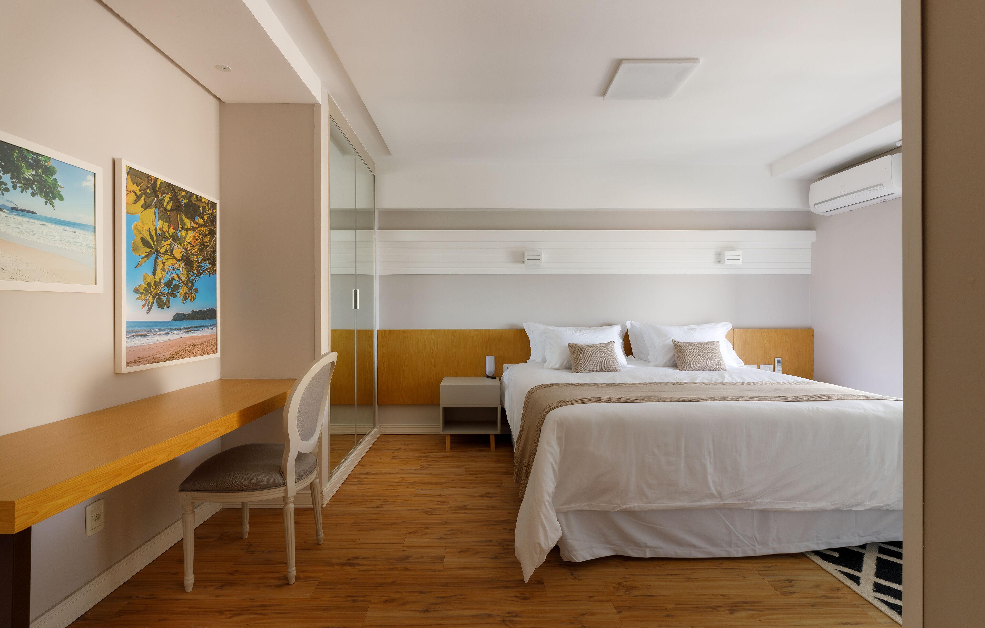 Quarto de hotel moderno com escrivaninha de madeira, cama com roupa de cama branca e duas pinturas na parede.