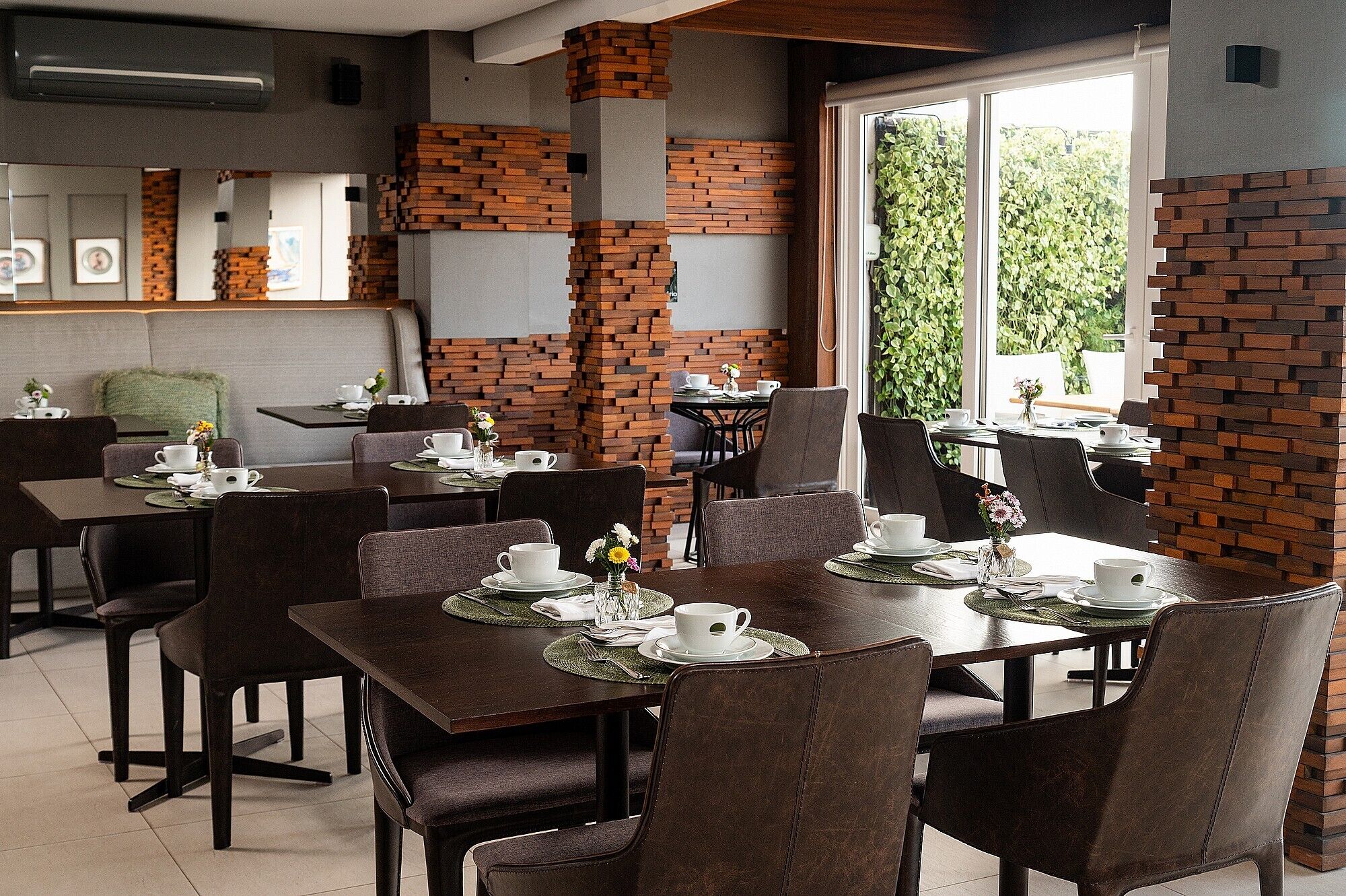 Zona pranzo moderna con dettagli in mattoni, diversi tavoli e sedie, e vista su piante verdi all'esterno.