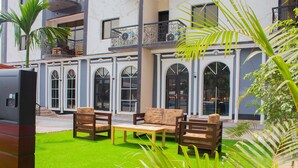 An outdoor seating area with a green lawn, wooden furniture, and a building with arched windows and balconies. - Marcsons Hotels and Resorts (Limbe)