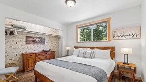Un dormitorio con cama de madera, mesita de noche, cómoda, banco, ventana con vista a árboles y pared decorada con un cuadro de un oso y un cartel que dice 'elige la amabilidad'.