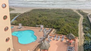 Une piscine sur le toit, dotée de chaises longues et d’un parasol en chaume, offrant une vue sur une plage et l’océan.