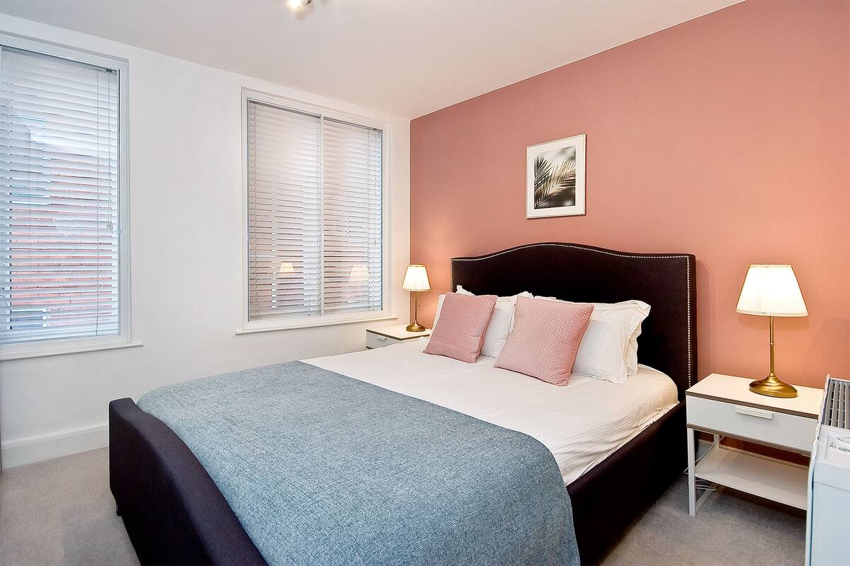 A bedroom with a bed, two bedside tables, a nightstand, a window with blinds, and a wall-mounted picture.