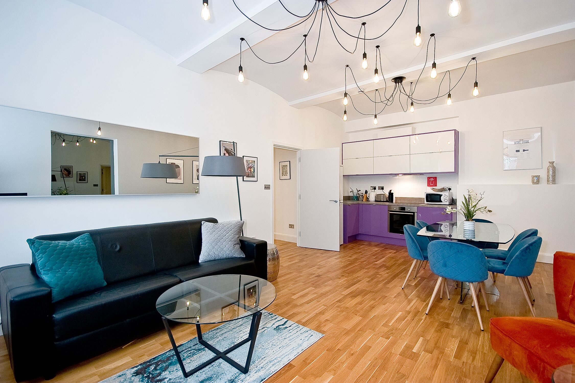 Una sala moderna con un sofá negro, una mesa de centro de vidrio y sillas azules. Se ve una cocina con armarios morados y una mesa de comedor al fondo.