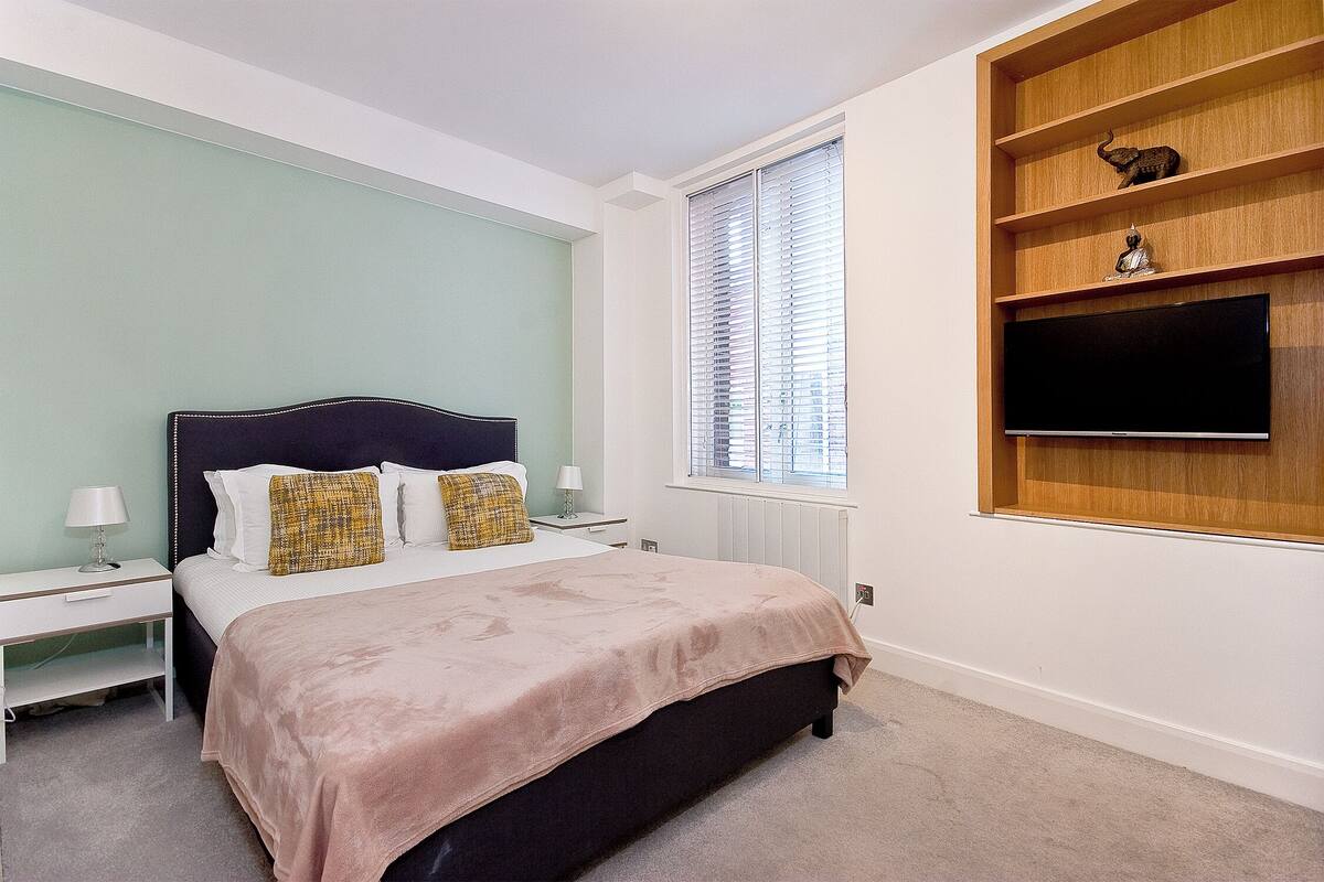 A bedroom with a bed, bedside tables, a window with blinds, and a built-in shelf with decorative items.