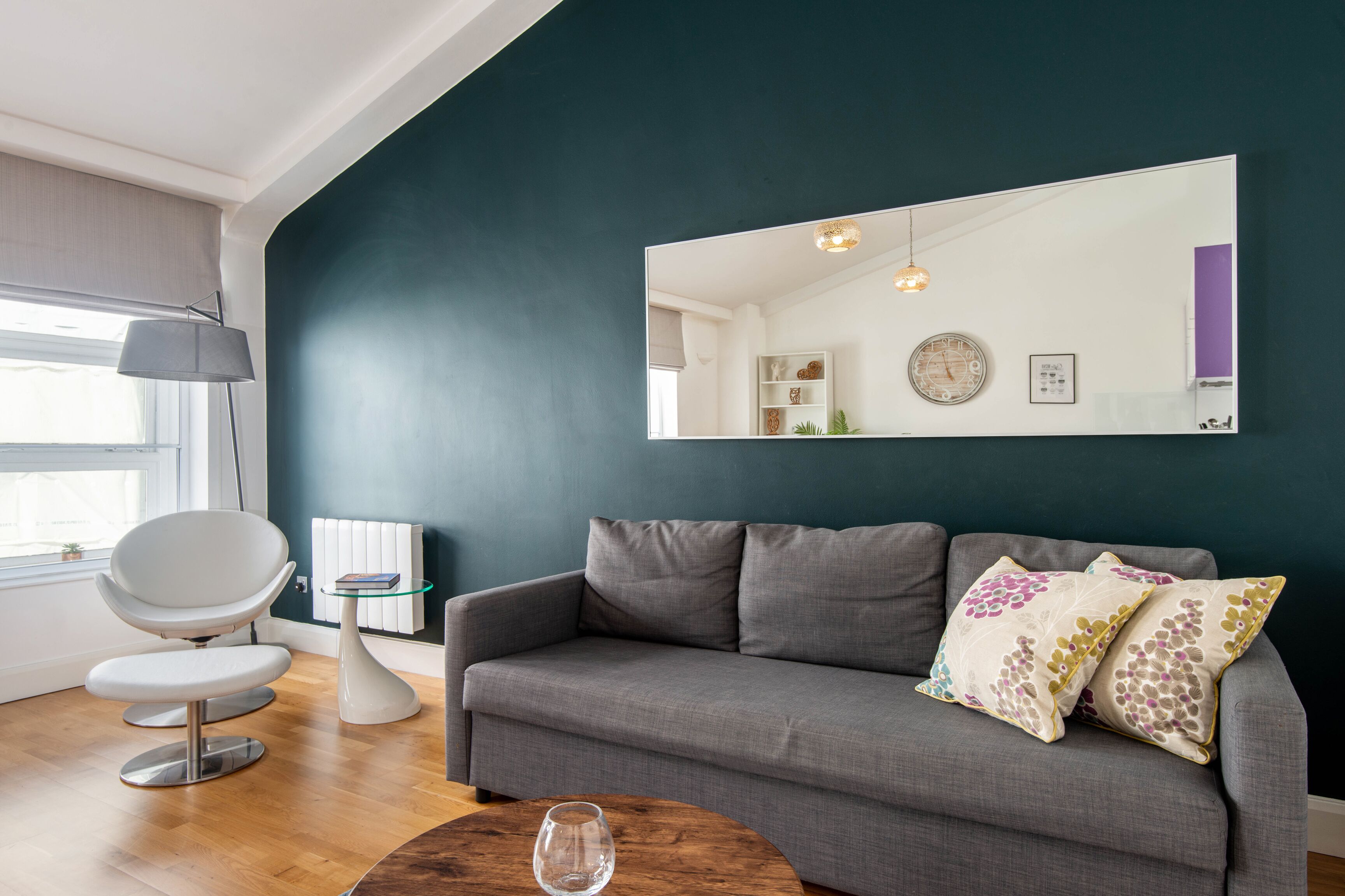 A modern living room with a grey sofa, a round coffee table, a white chair, and a large mirror on the wall.