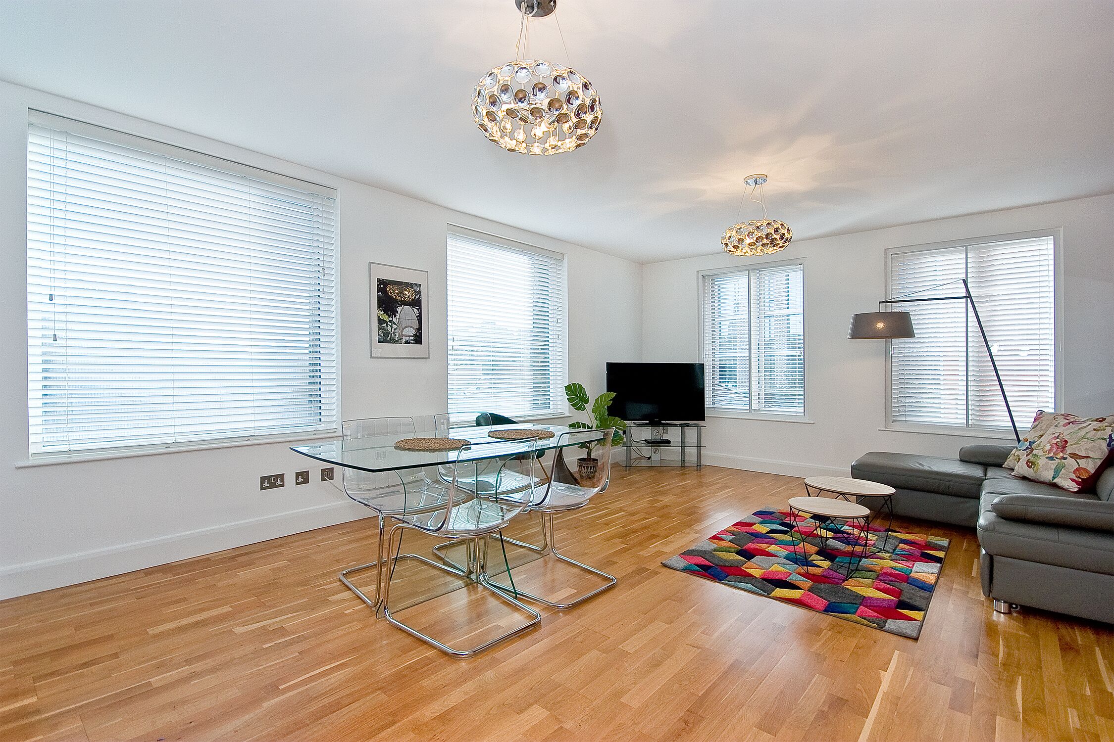 Een moderne woonkamer met een glazen eettafel, een grijze bank en een kleurrijk vloerkleed.