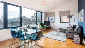 A modern living room with a glass dining table, green chairs, a grey sofa, and a city view through large windows. - Mulberry House London by City Living London (London)