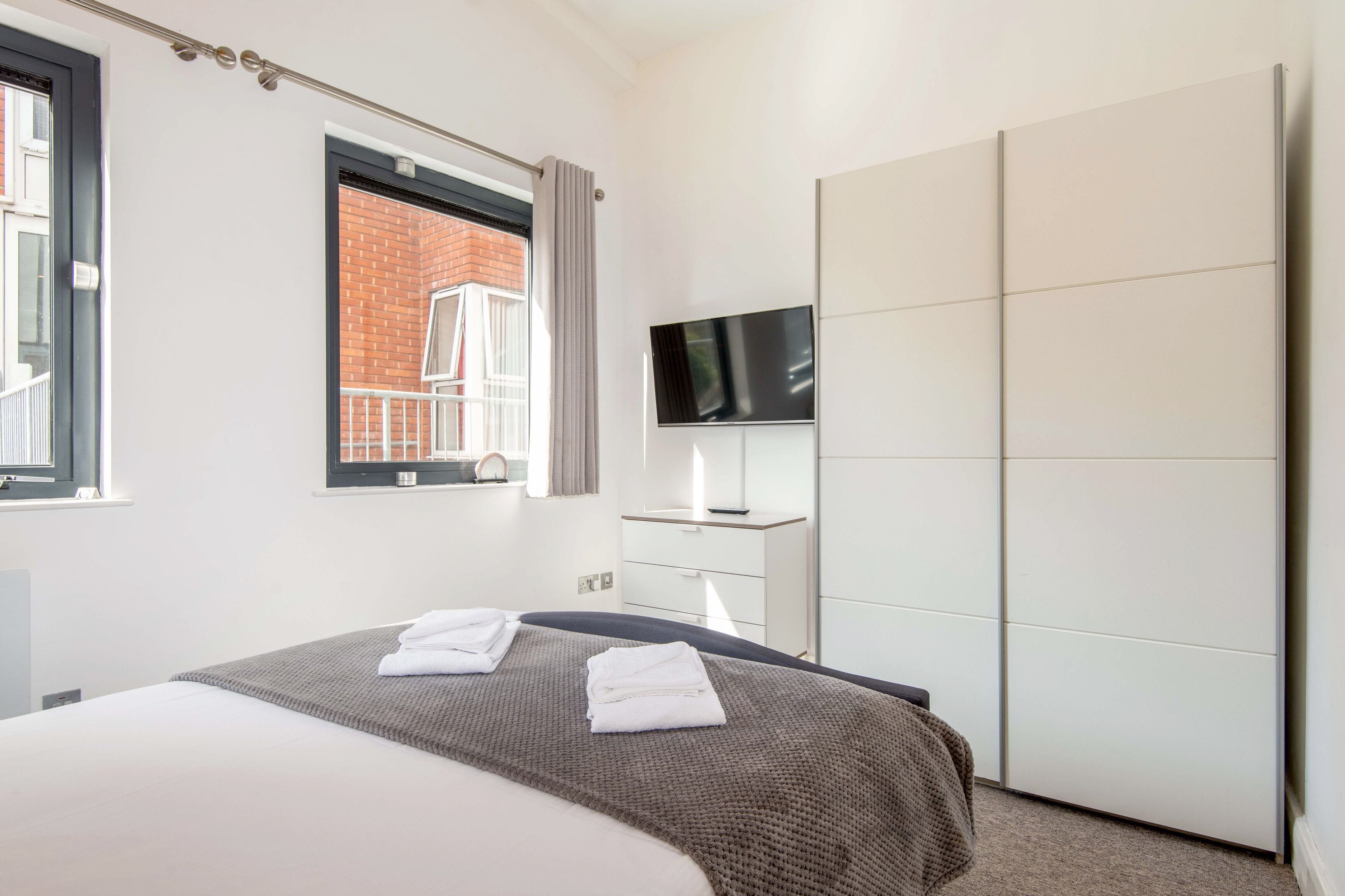 A hotel room with a bed, a TV mounted on the wall, a wardrobe, and a window with a view of a brick building.