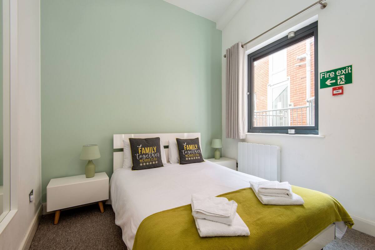 A neatly made bed with two pillows, a green throw blanket, and folded towels. There is a nightstand with a lamp, a fire exit sign, and a window with a view of a building.