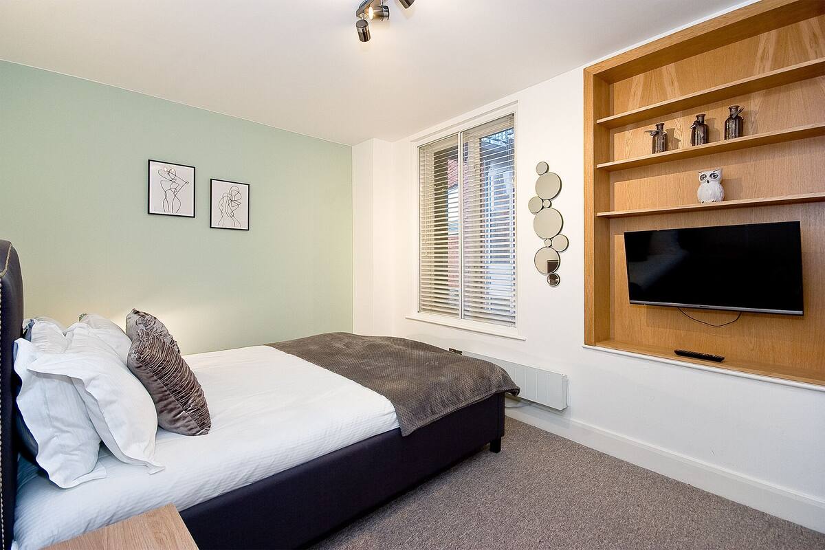 A bedroom with a bed, a TV, a window with blinds, and wall-mounted shelves.