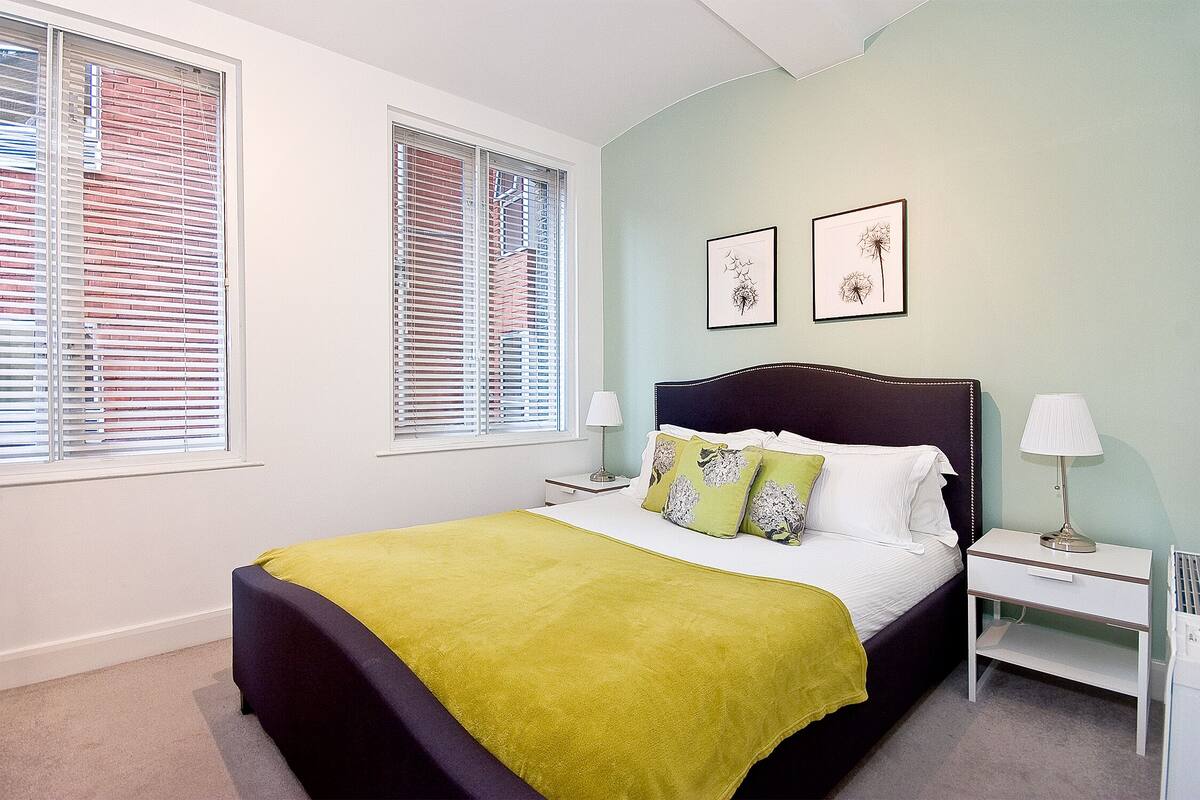 A bedroom with a bed, two bedside tables, a nightstand, and two windows with blinds.