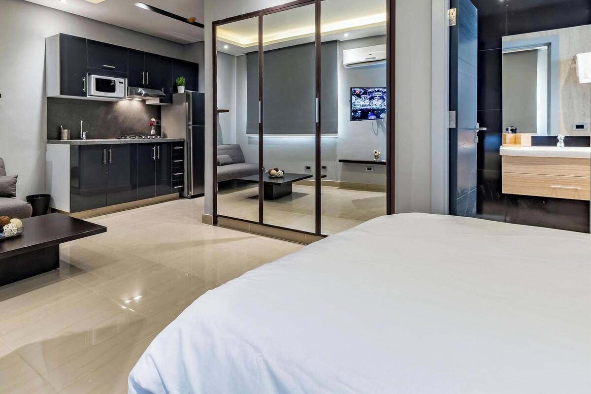 A modern hotel room with a sofa, a bed, a coffee table, a TV, a kitchen area, and a sliding door leading to a bathroom.