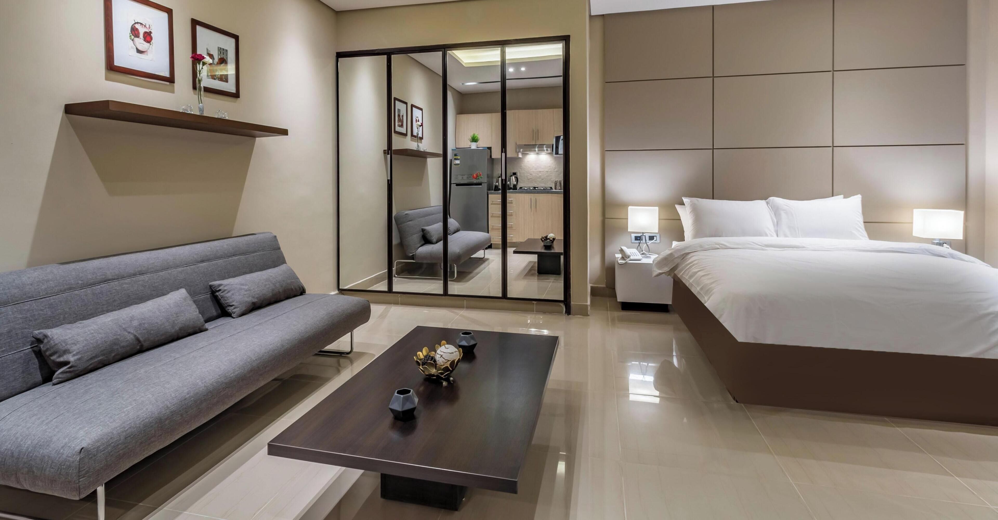 A modern hotel room with a sofa, a bed, a coffee table, and a sliding door leading to a kitchen area.