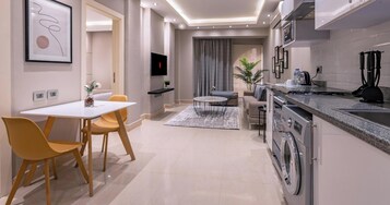 A modern kitchen with a built-in washer and dryer, a dining area with a white table and yellow chairs, and a living area with a sofa and a coffee table.