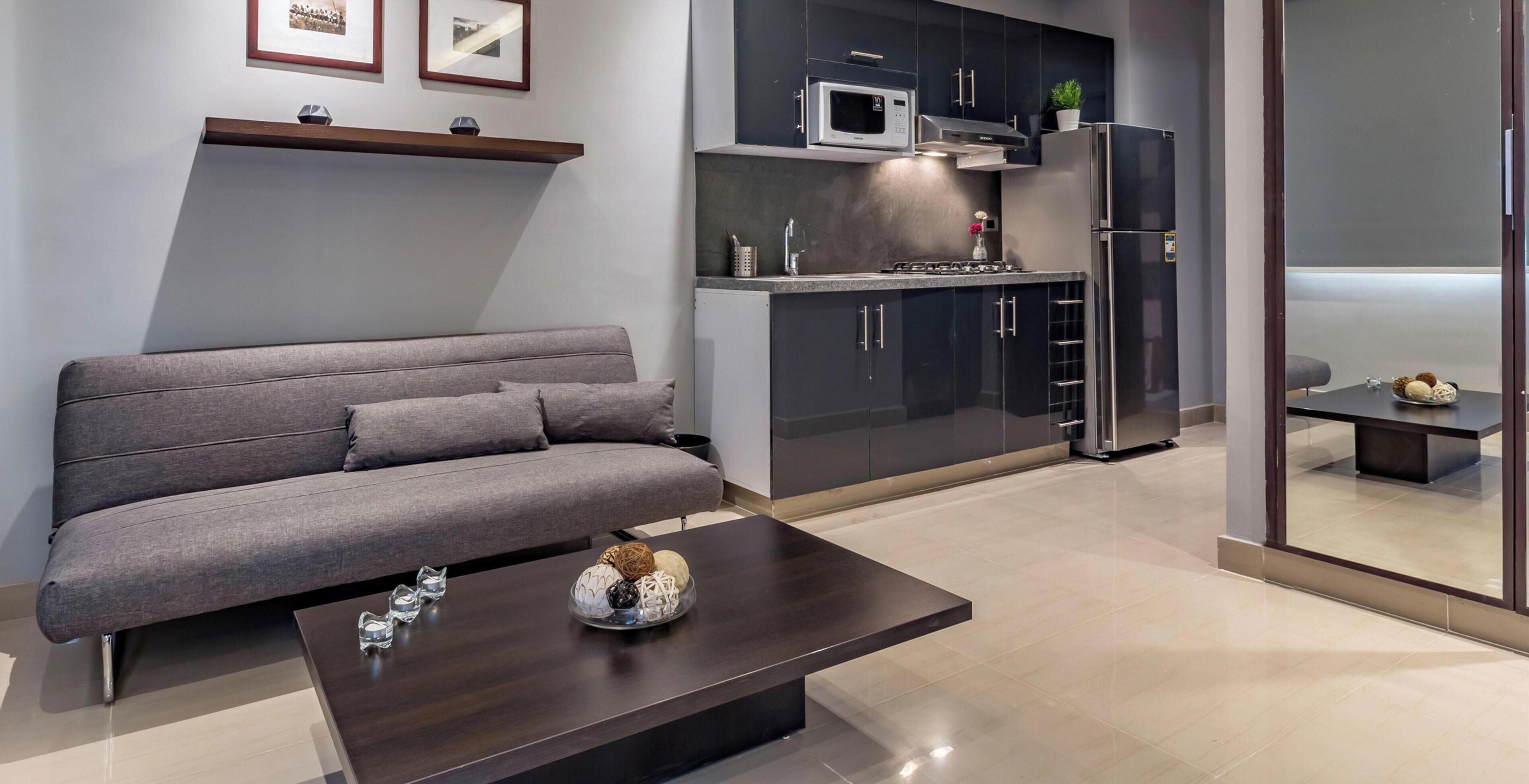 A modern living room with a grey sofa, a dark coffee table, and a kitchen area with dark cabinets, a microwave, and a refrigerator.