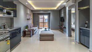 A modern living room with a sofa, ottoman, and a TV. There is a kitchen area with a stove and microwave. The room has a large window and a balcony. - Prime Residence New Cairo (New Cairo)
