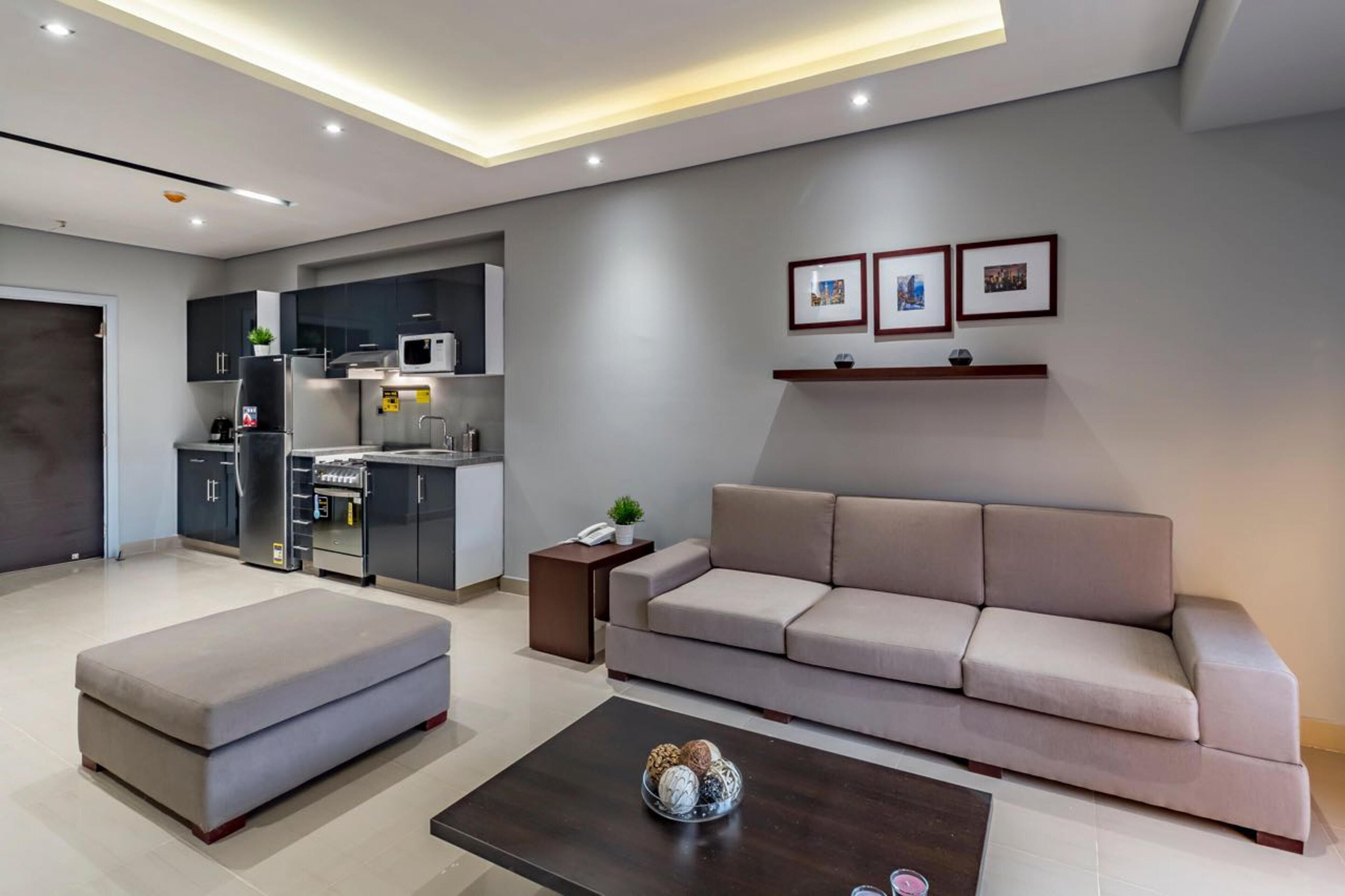Uma sala de estar moderna com sofá, puf e mesa de centro, área de cozinha com eletrodomésticos e quadros nas paredes.