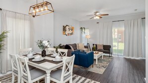 Un salon spacieux comprenant un coin repas, un ventilateur de plafond et une grande fenêtre avec des rideaux.
