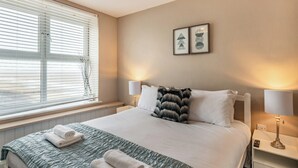 A neatly made bed with a green quilt, two white pillows, and a fur throw. There are two bedside lamps, a framed picture on the wall, and a window with blinds.