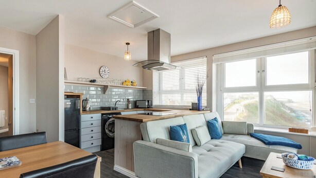 A modern kitchen with a central island, a dining area with a wooden table and chairs, and a living area with a sofa and a view of the outdoors.