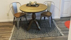Una mesa redonda de madera con dos sillas de metal, un letrero decorativo en la pared y una alfombra estampada.