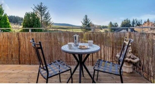 Eine Holzdar Terrasse mit einem runden Tisch und zwei Stühlen, einem Metallzaun und Blick auf eine ländliche Landschaft.