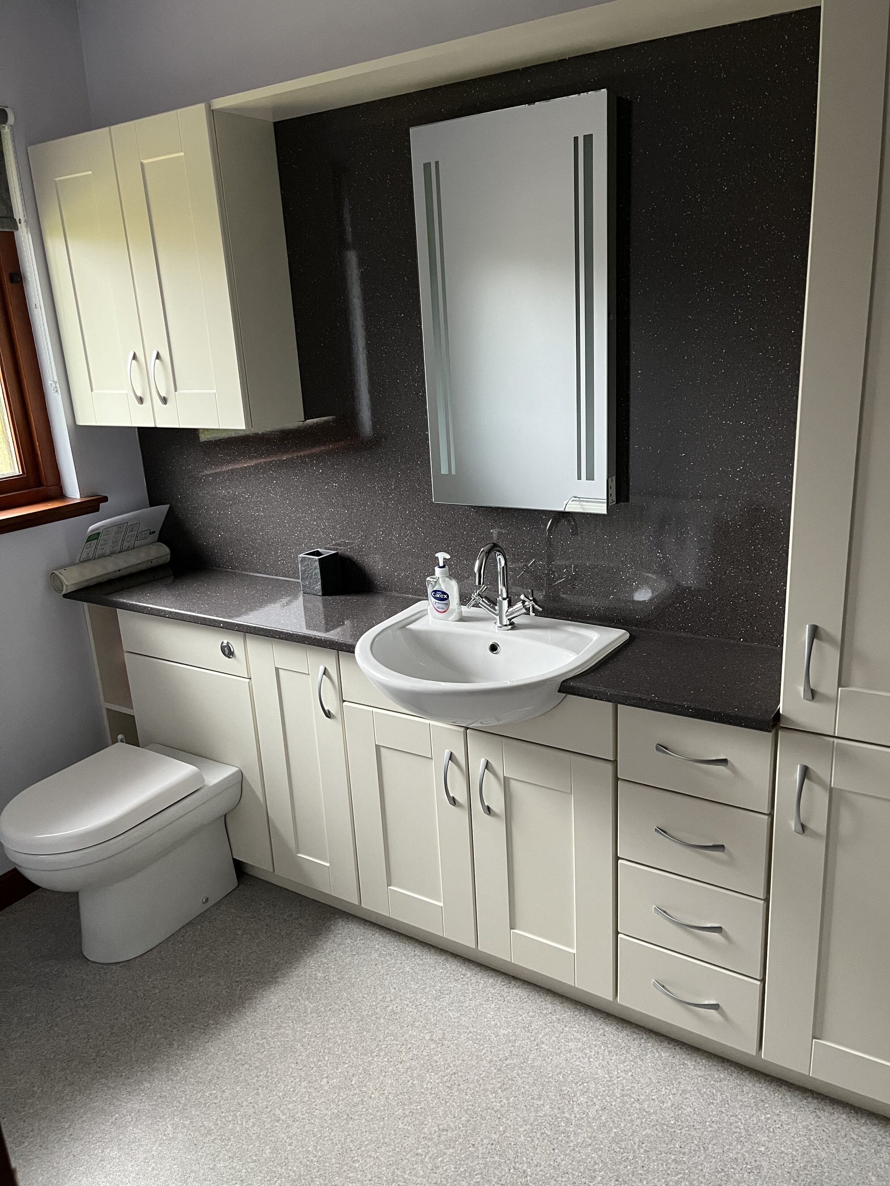 Une salle de bain moderne avec des toilettes blanches, un lavabo surmonté d’un miroir et des meubles dotés de poignées argentées.