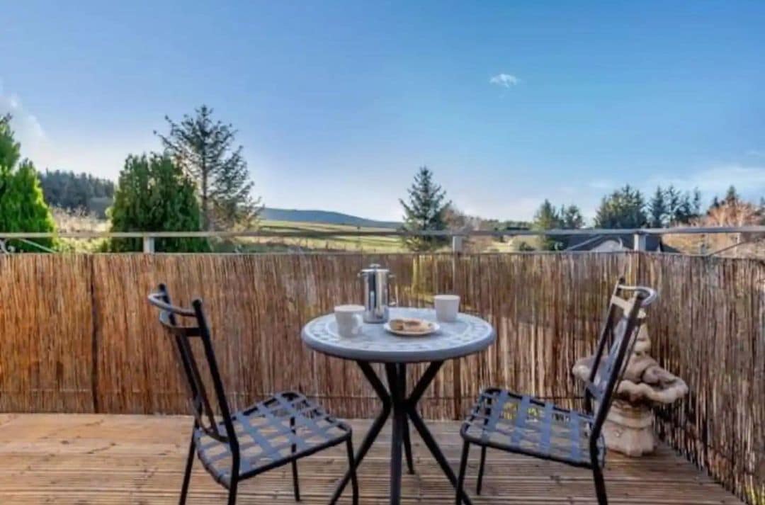 A wooden deck with a round table and two chairs, a metal fence, and a view of a rural landscape.