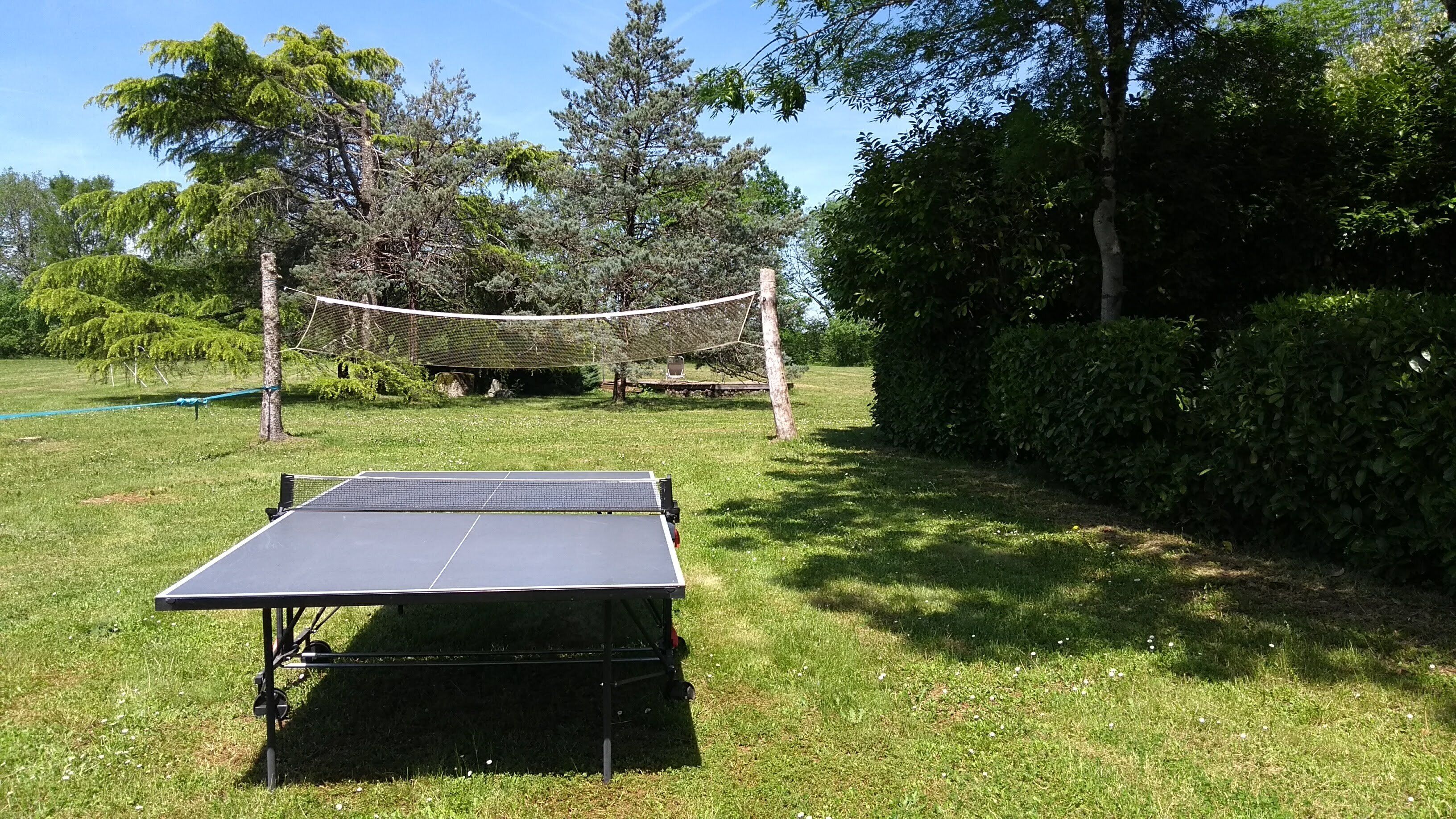Una mesa de ping pong portátil en un espacio al aire libre con césped, con una instalación de voleibol con red al fondo.