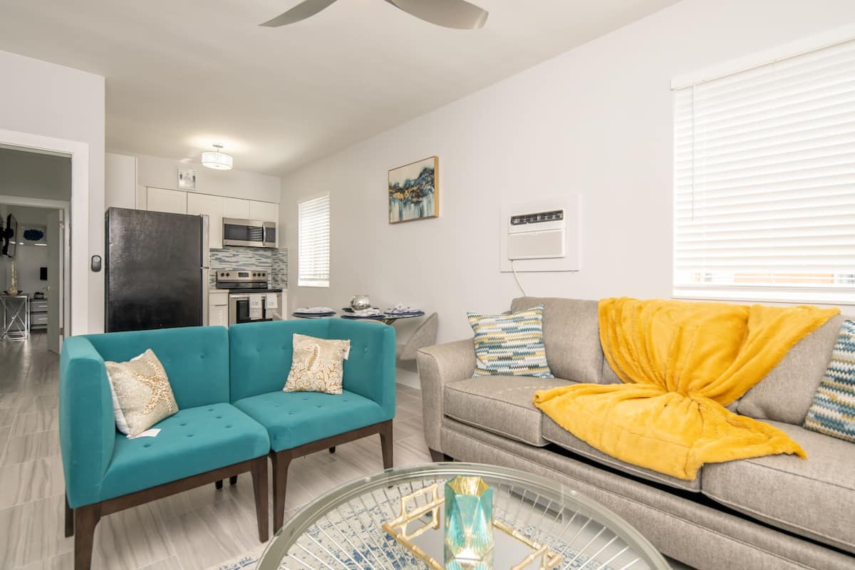 A modern living room with a teal sofa, a grey sofa with a yellow throw, and a glass coffee table. There is a kitchen in the background with white cabinets and a microwave.