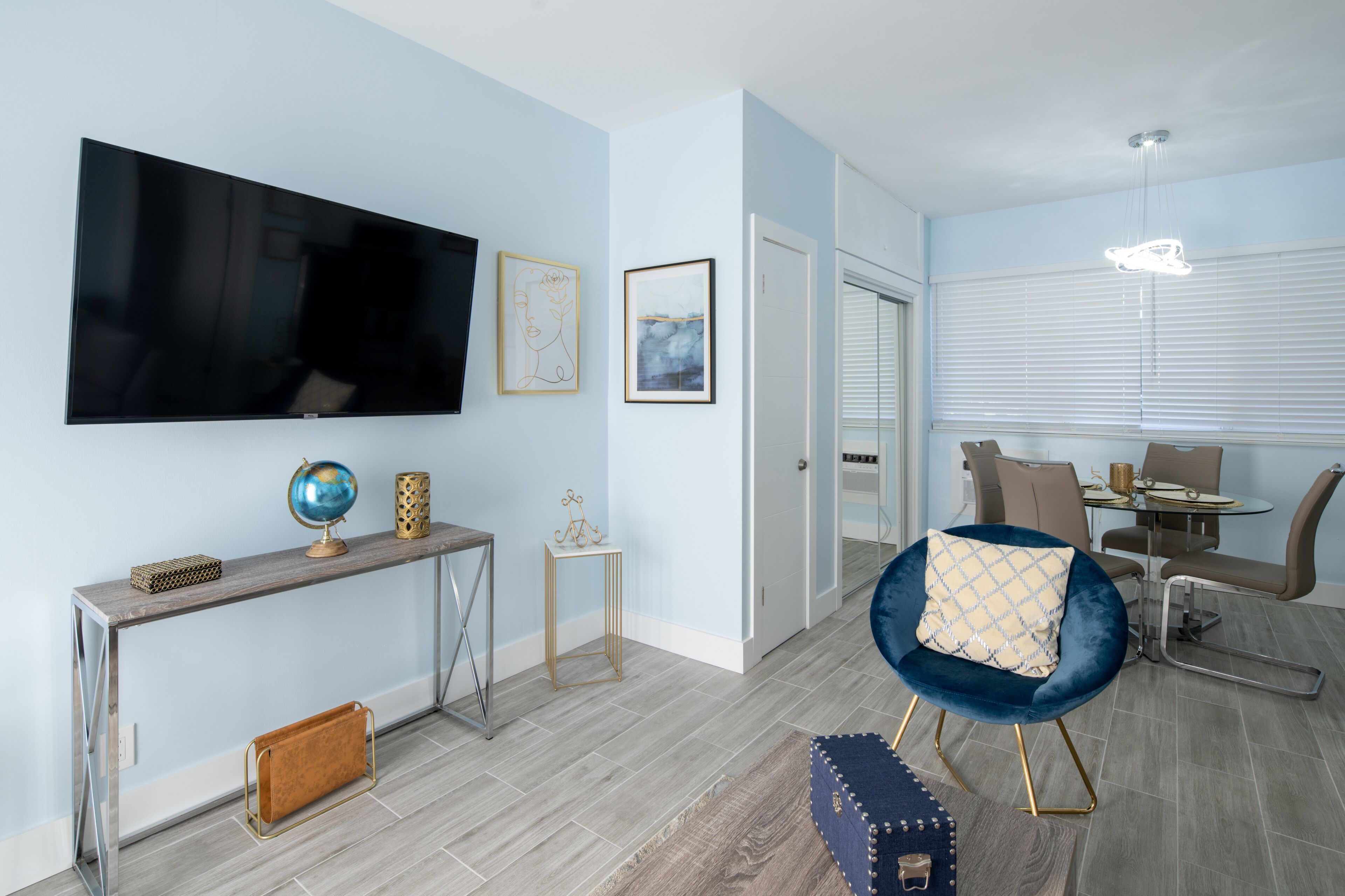 A modern living room with a flat-screen TV, a console table with decorative items, a blue armchair, and a dining area with a table and chairs.