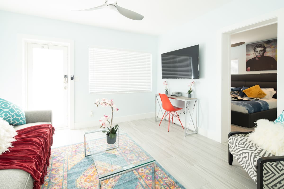 A modern living room with a sofa, a coffee table, a desk with a chair, a bed, and a TV mounted on the wall.