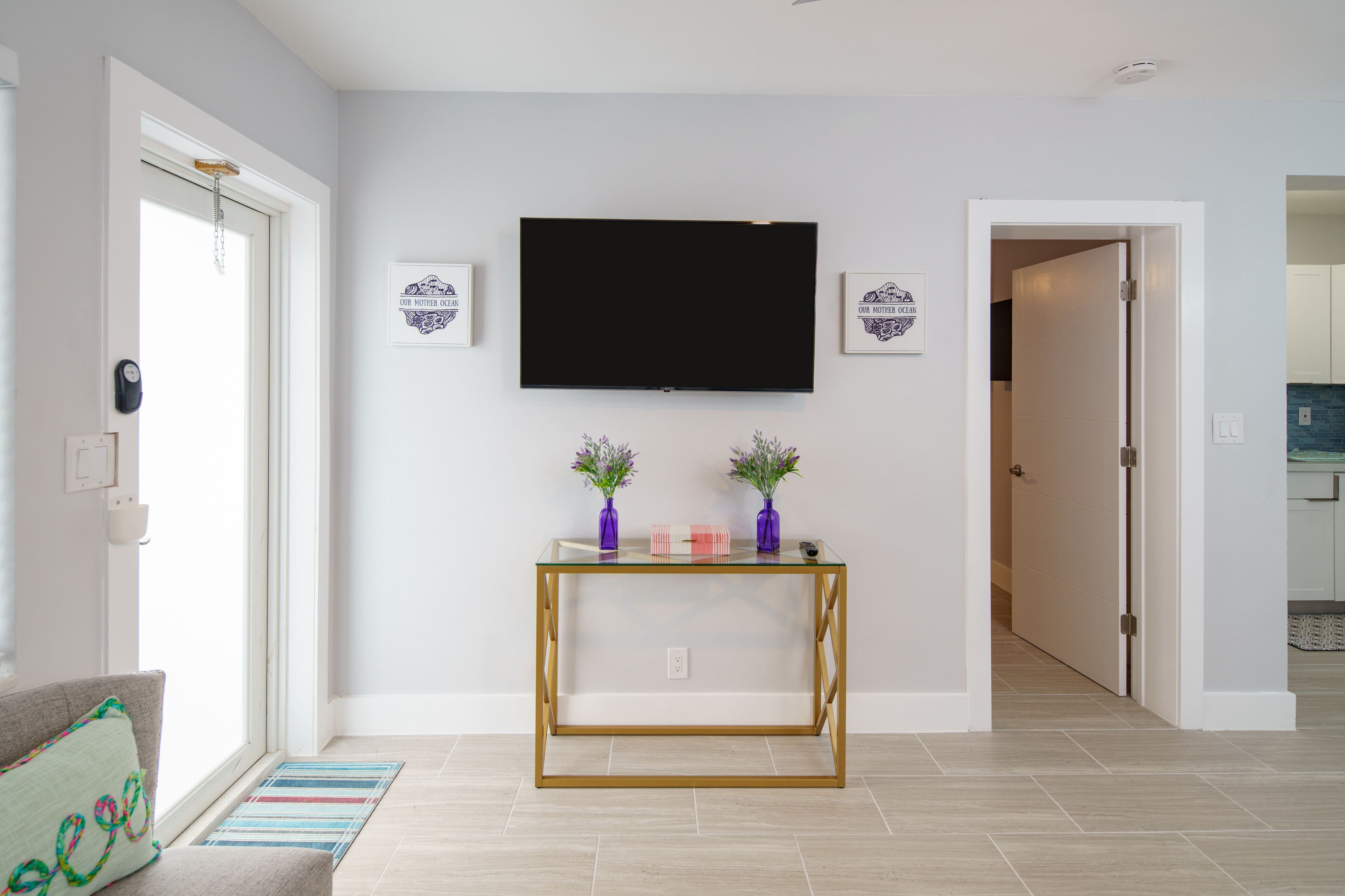 Una sala de estar moderna con un televisor de pantalla plana montado en la pared, una mesa de centro de vidrio con objetos decorativos y una puerta que da a otra habitación.