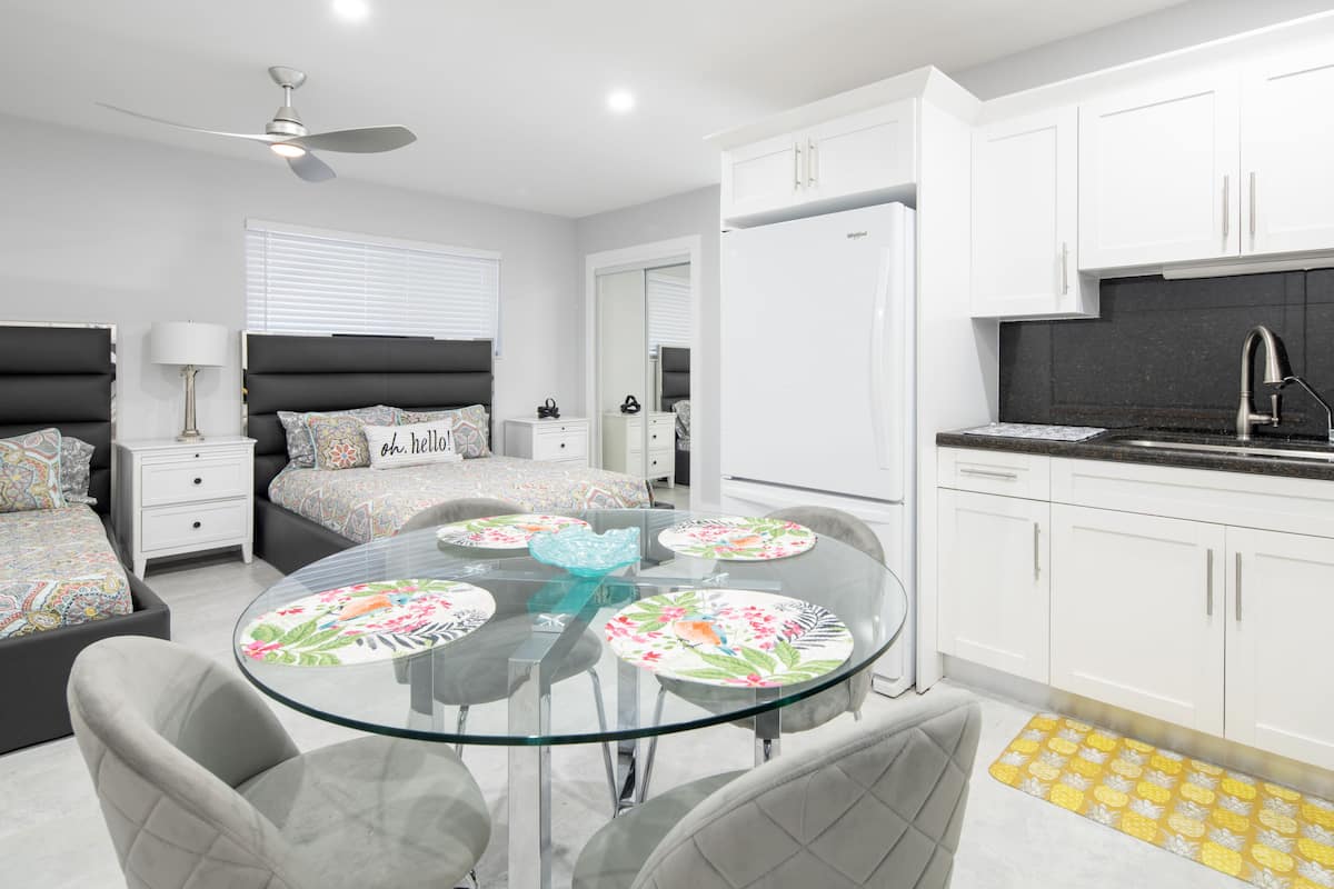 A modern bedroom with a glass dining table, a bed with patterned bedding, a white wardrobe, and a kitchen area with a black countertop.