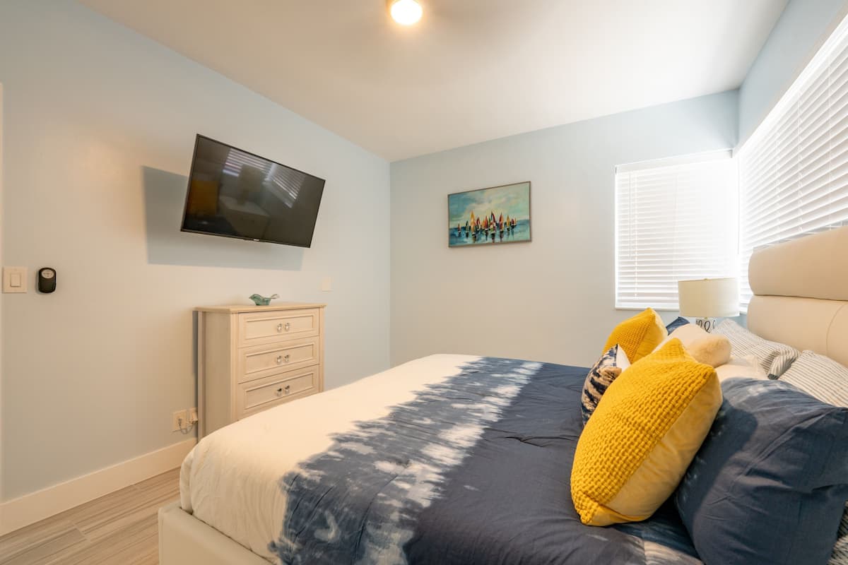 A bedroom with a bed, a television mounted on the wall, a dresser, and a window with blinds.