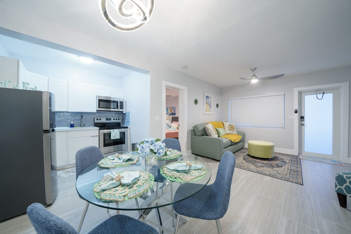 A modern kitchen with white cabinets, a microwave, and a refrigerator. There is a dining area with a glass table and blue chairs. A gray sofa and a yellow ottoman are in the living room.