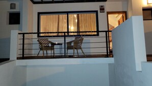 Varanda com duas cadeiras de vime e uma pequena mesa, janela com cortinas e um corrimão.