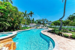 Une piscine à bord incurvé, entourée d’une clôture blanche en bois, de palmiers et d’une maison en arrière-plan.