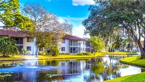 A building with a red roof, surrounded by a well-maintained lawn and a pond with a fountain. - *Casa Bella* Waterfront View! 2BR, w/2 Pools, Mins to IMG/Beach/Anna Maria Isl (Bradenton)
