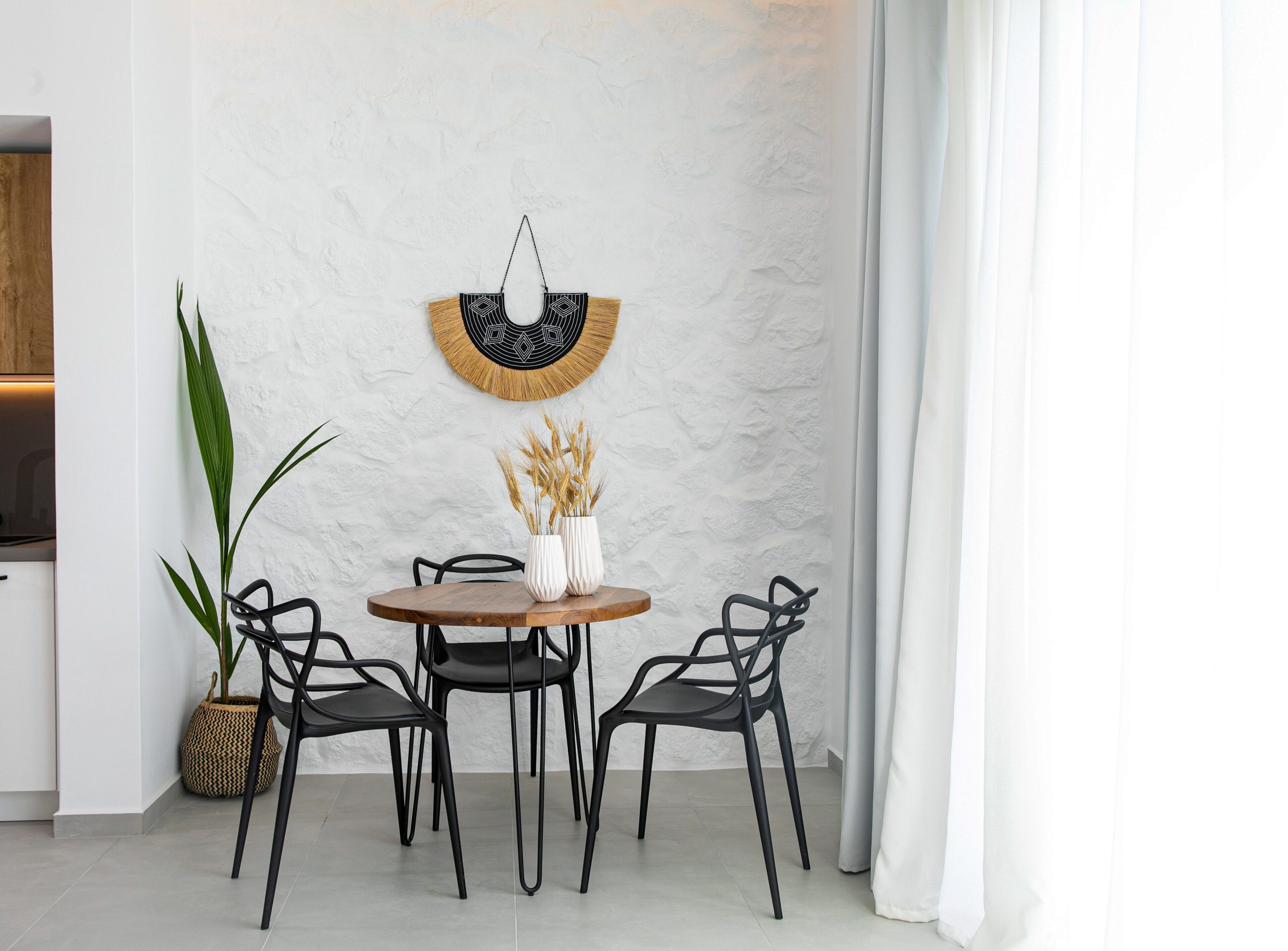 Un coin repas avec une table ronde en bois, quatre chaises noires, un vase blanc contenant des plantes séchées et un mur texturé agrémenté d’un élément décoratif suspendu.